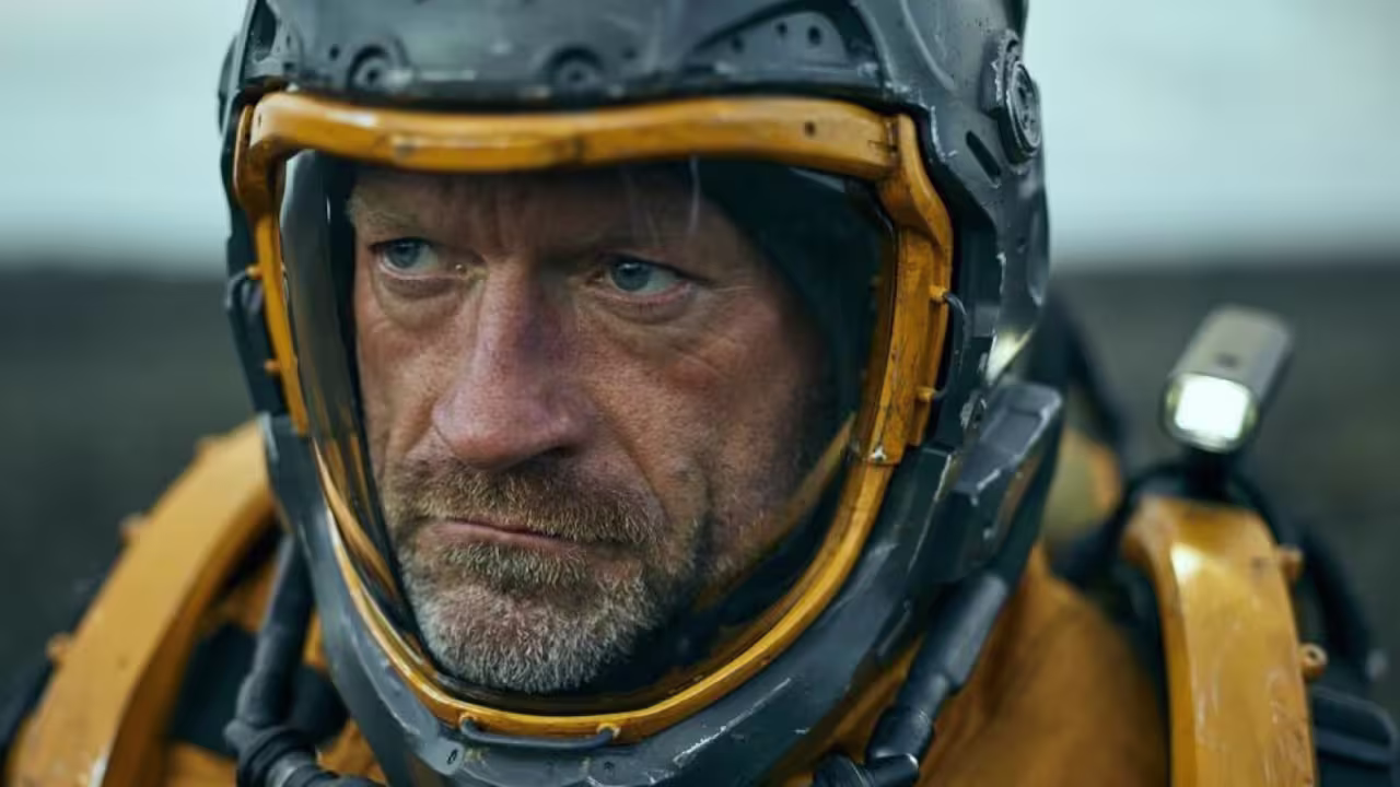 Close-up of a man with a beard wearing a weathered yellow and gray sci-fi helmet and suit, looking seriously to the side.