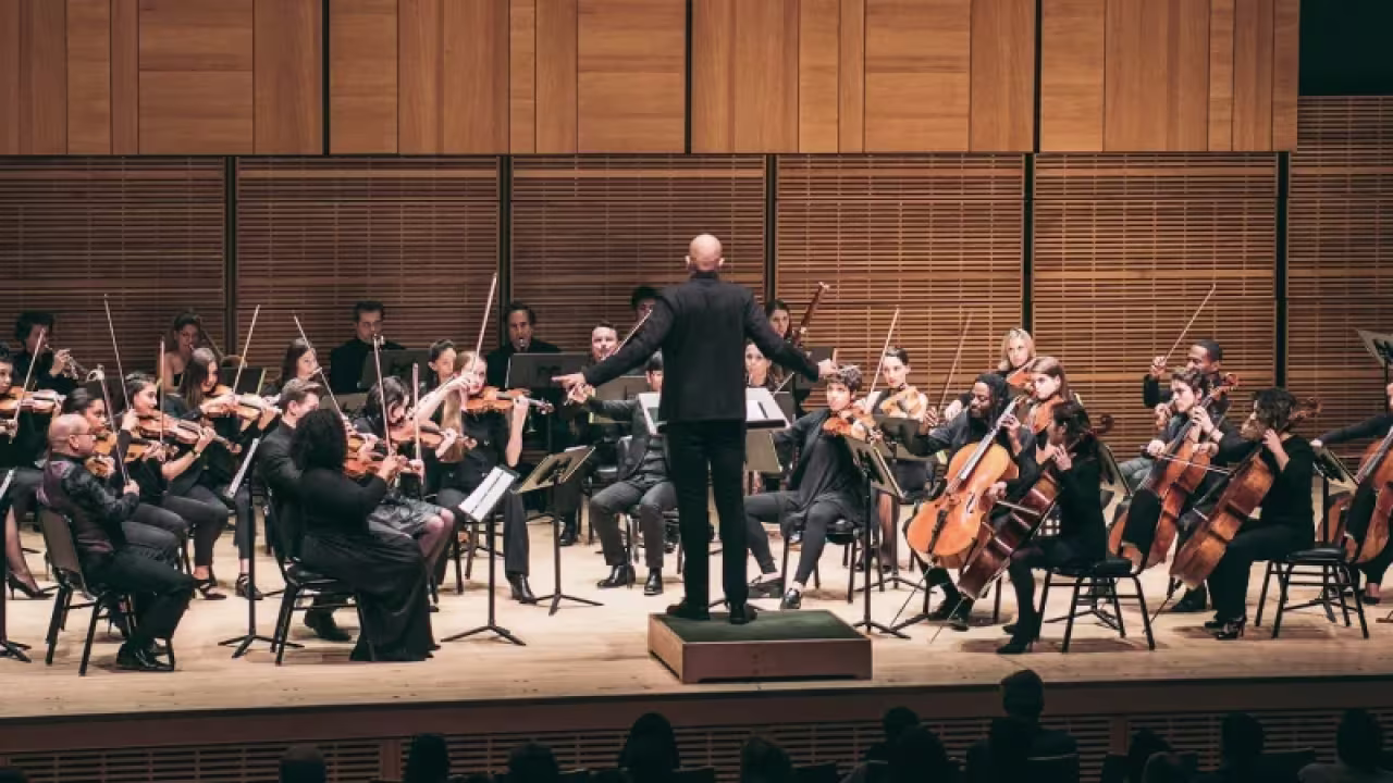 Conductor leading a symphony orchestra with string and woodwind musicians on stage in a concert hall.