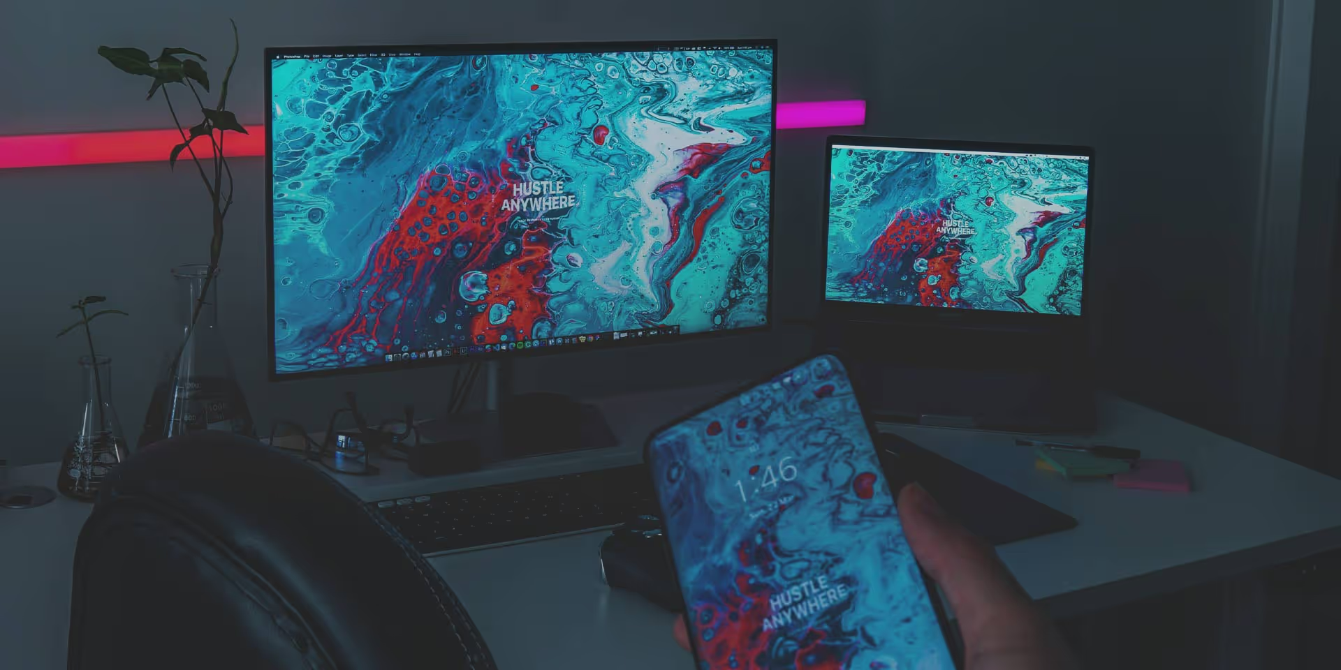 A workspace with a monitor, laptop, and smartphone all displaying a colorful abstract background with the text &"HUSTLE ANYWHERE".