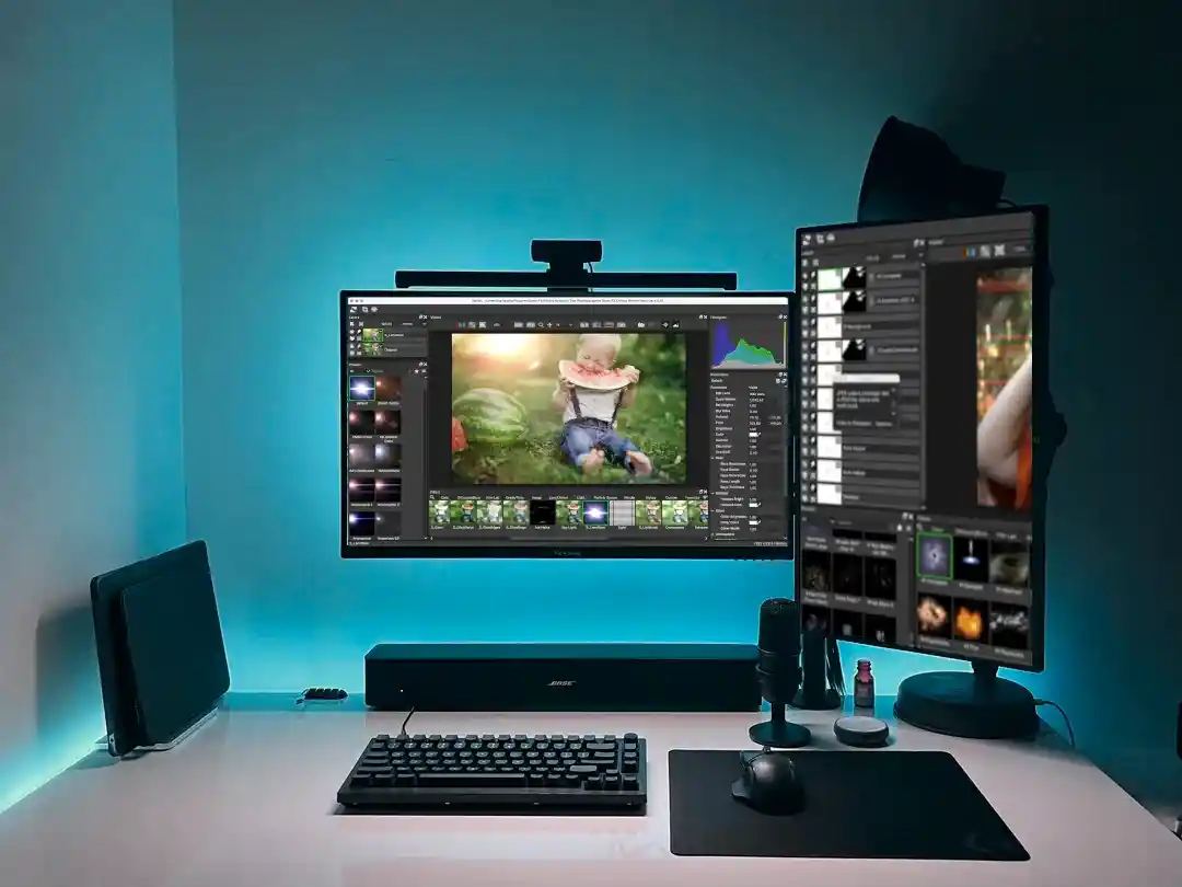 Cyan-lit desk with a landscape and a portrait monitor running Optics, editing a photo of a child sitting in a garden.