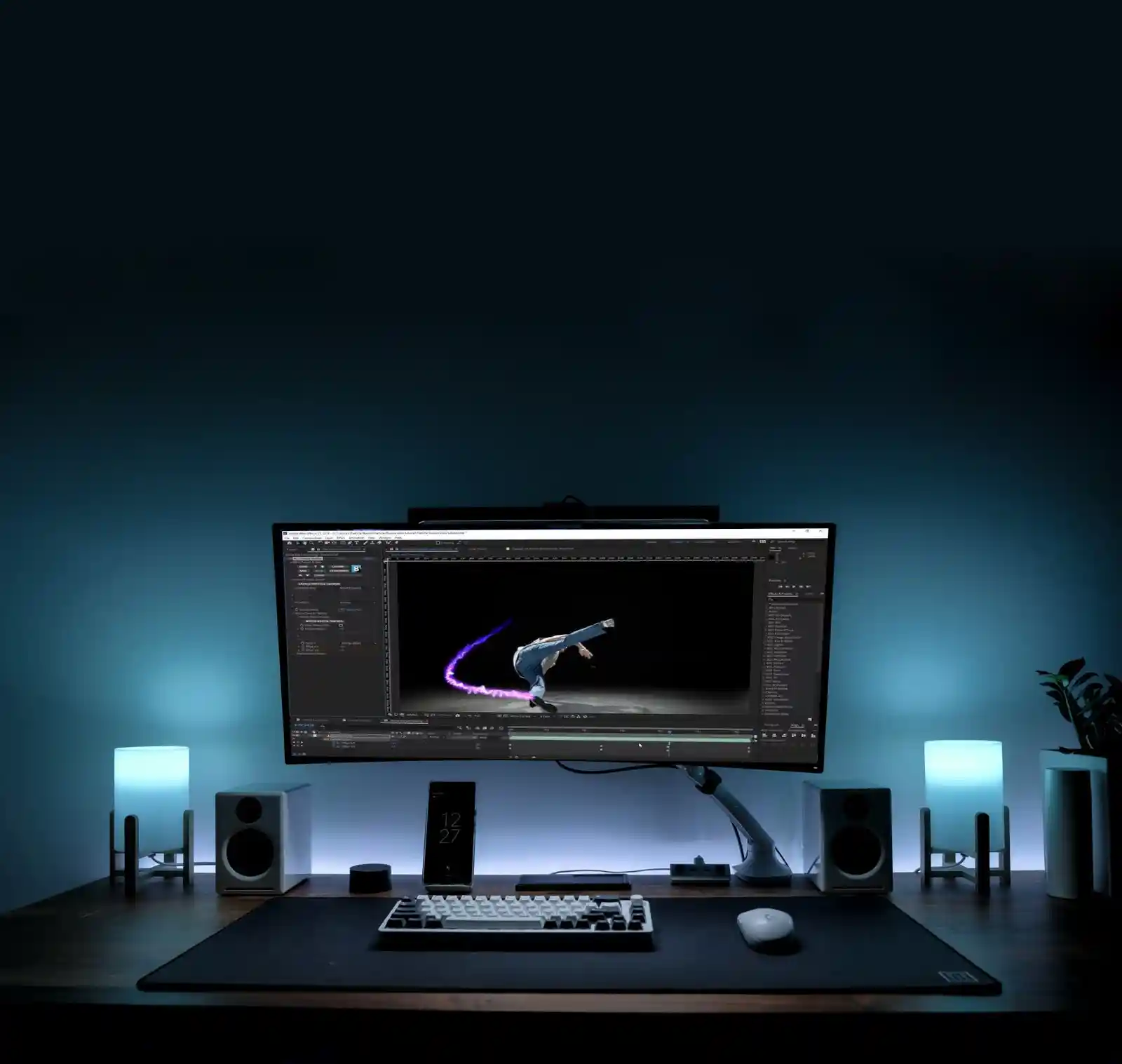 Blue-lit desk with a wide monitor running Particle Illusion, showing a dancer trailed by purple light-streak particles, between glowing lamps, speakers and a plant.